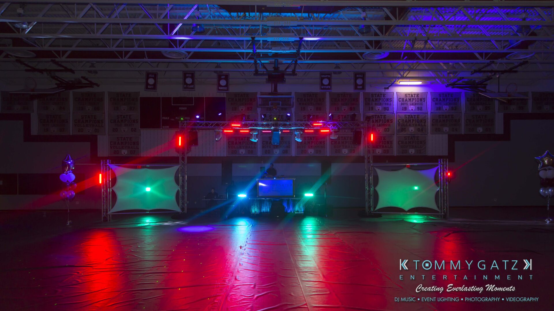 Empty gymnasium decorated for an event with colorful stage lights and two projector screens, branding for "Tommy Gatz Entertainment" visible at the bottom right.