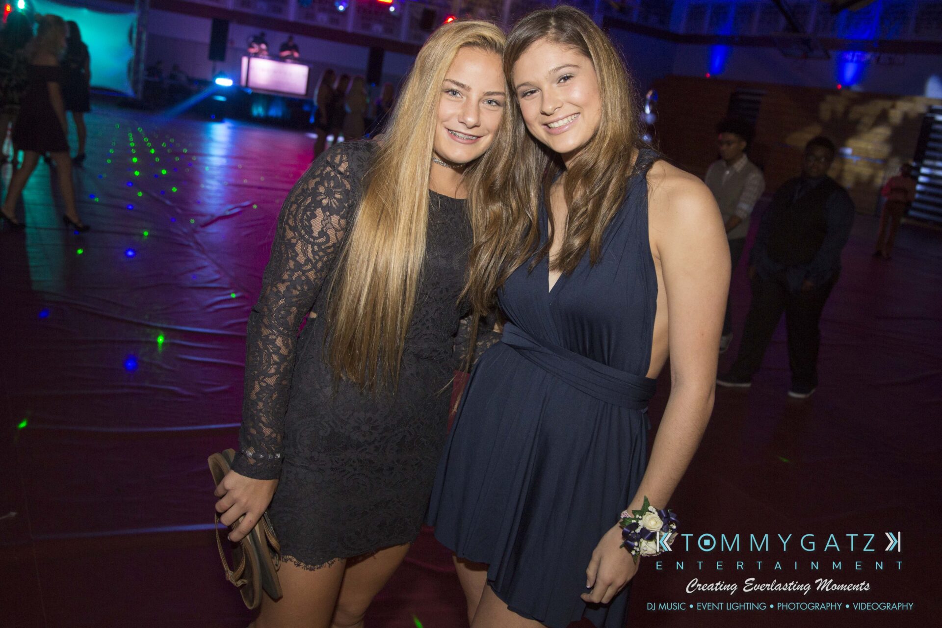 Two women in semi-formal attire smile at the camera in a dimly-lit event space with scattered colored lights.