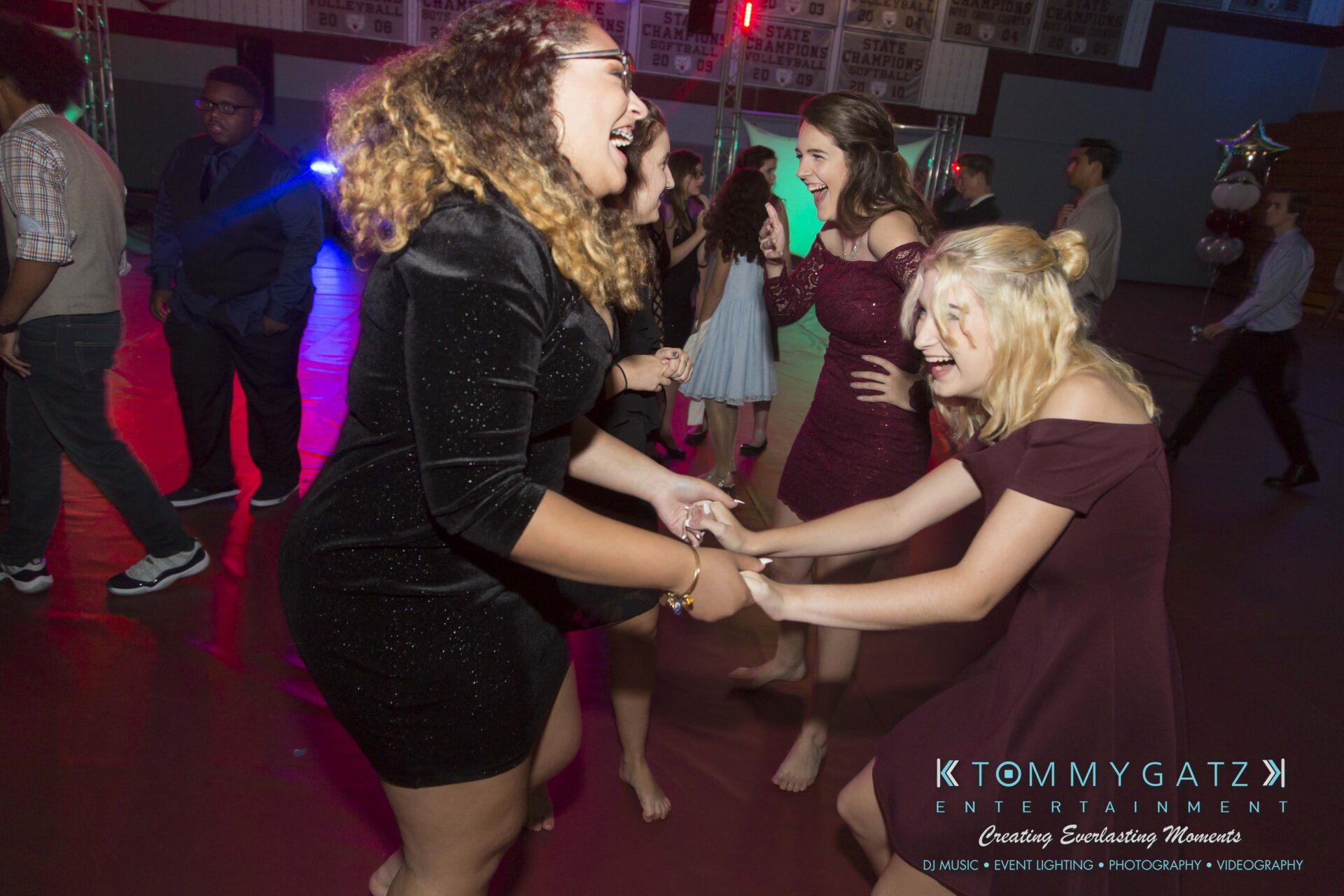 Three women in dresses are laughing and holding hands on a dance floor, with party lights and other people in the background.