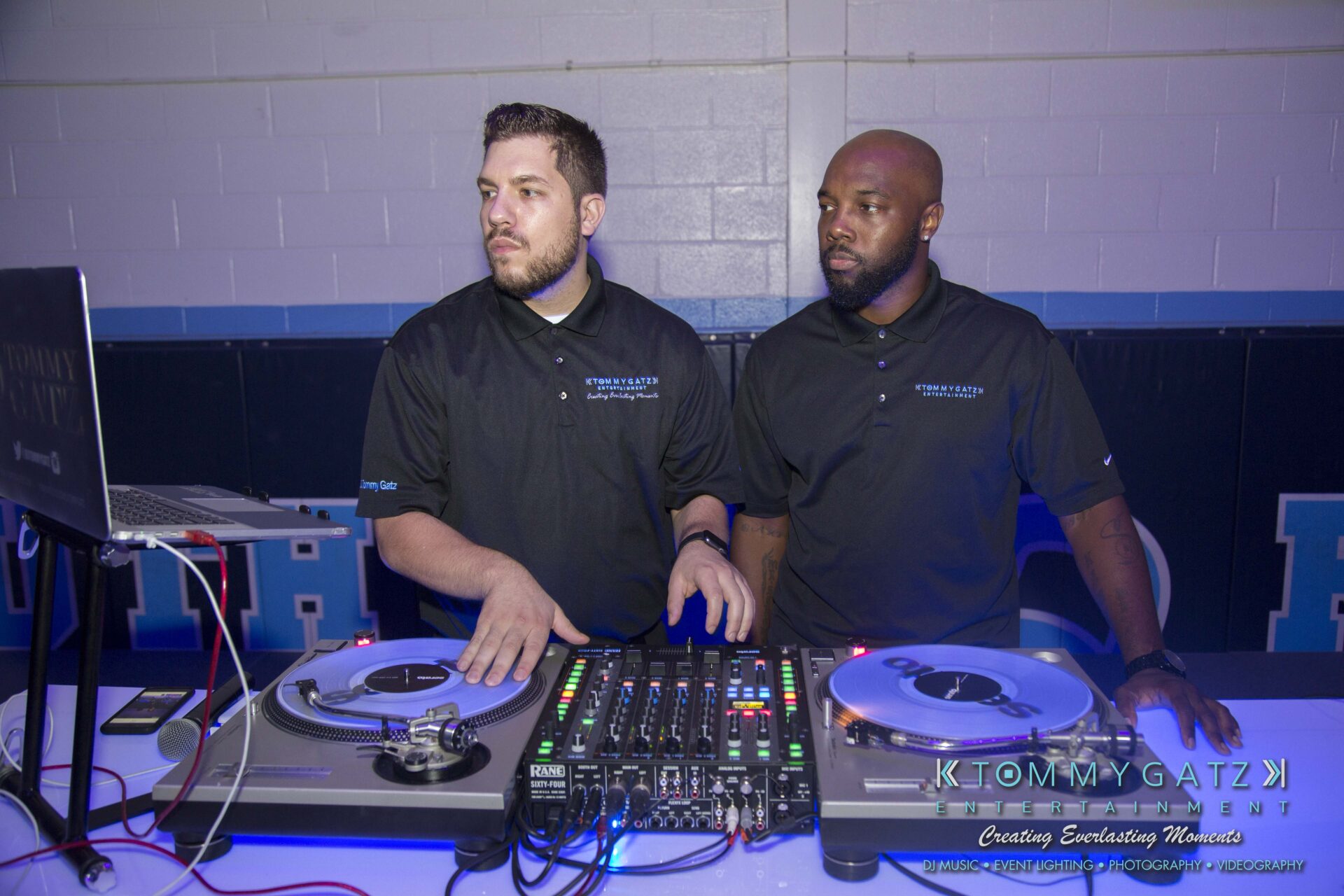 Two DJs in black shirts operate turntables and a mixer on a table in a room with blue accents, focused on equipment and surroundings.