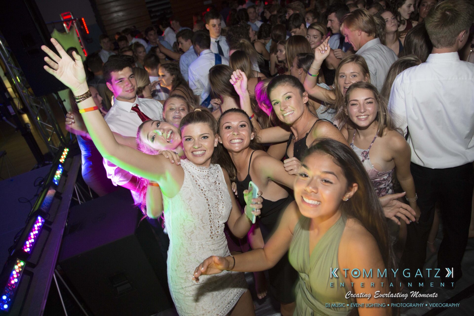A group of young people, dressed in formal attire, are gathered closely together at a lively event, some smiling at the camera. Colorful lights illuminate the scene.