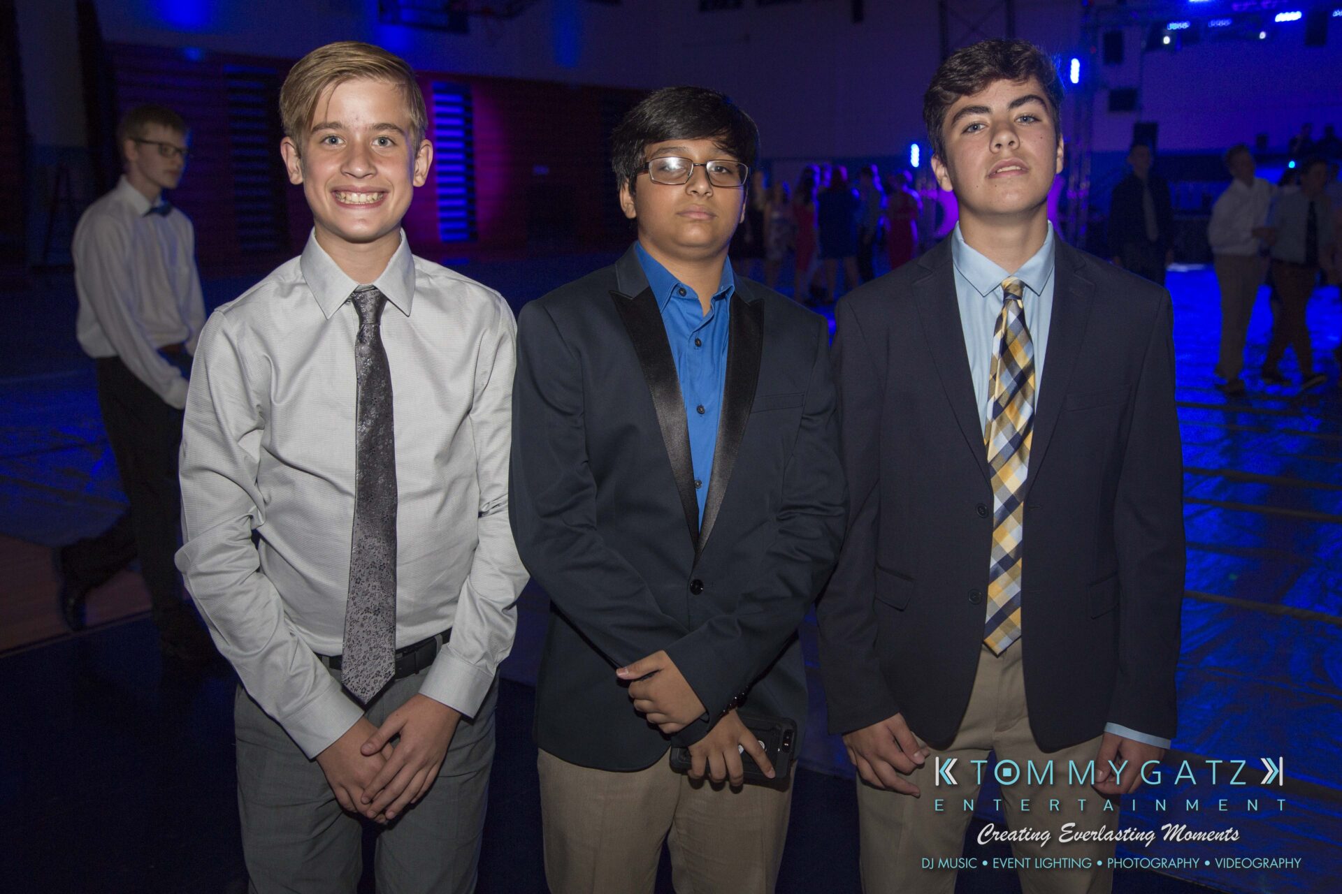 Three young individuals in formal attire pose for a photo in a dimly lit event space with blue lighting.