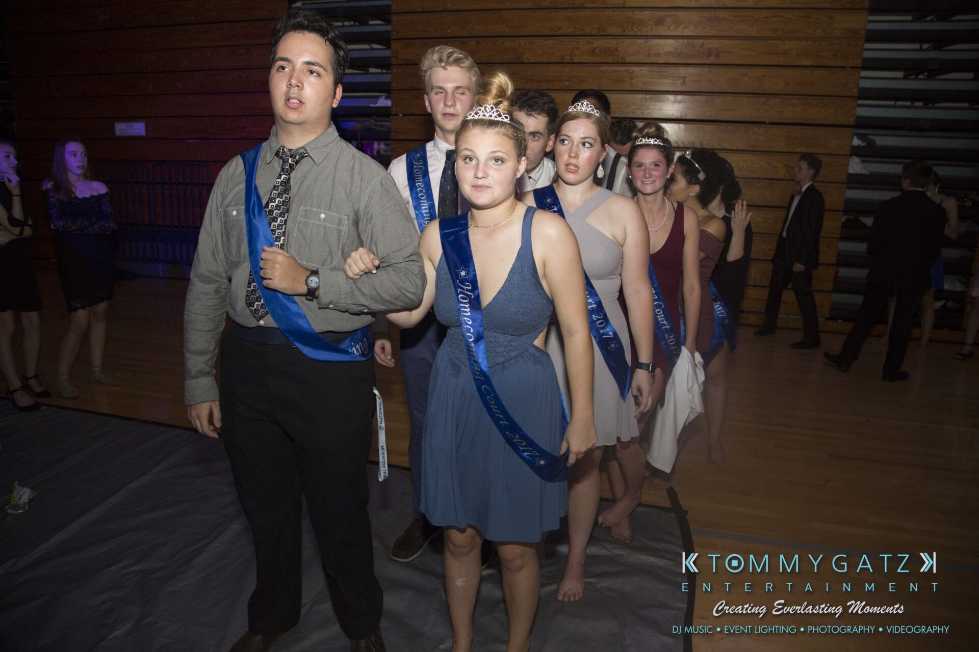 A group of young people in formal attire and tiaras line up in a gymnasium, holding blue sashes.