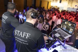 Two DJs in "Staff" shirts perform at a lively indoor event with a crowd dancing in the background.