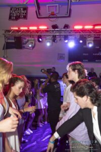 People dancing at an indoor event, with bright lights and an energetic atmosphere. A person in a black staff shirt is at the center. Signs for "STATE CHAMPIONS BASEBALL 2013" are visible.