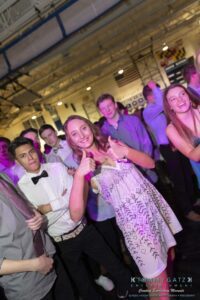 A group of young people dancing at an indoor event, with one girl in the center giving a thumbs-up. Background shows dim lighting and event decorations.