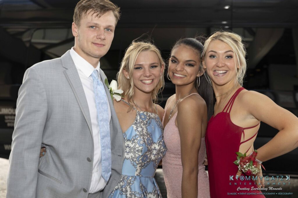 Four young adults dressed in formal attire pose together, smiling, with a vehicle in the background.