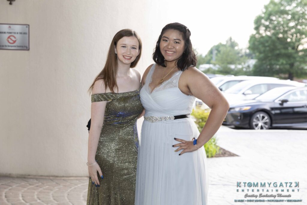 Two women in formal dresses pose outdoors on a sunny day. A car and greenery are visible in the background.