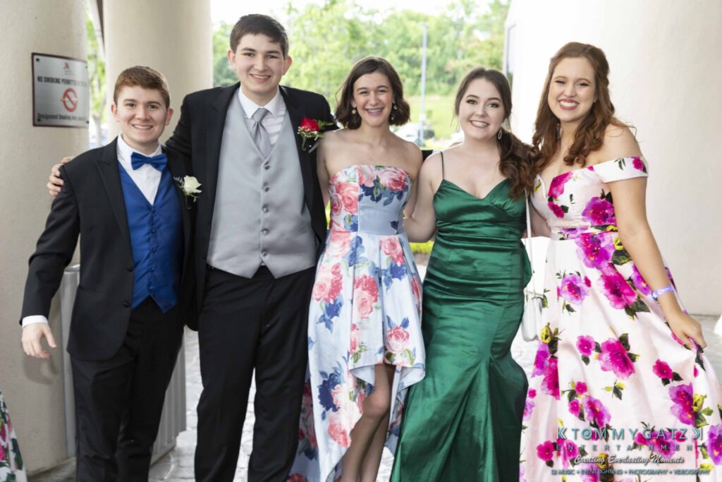 A group of four women and one man, dressed in formal attire, stand together smiling in an outdoor setting.