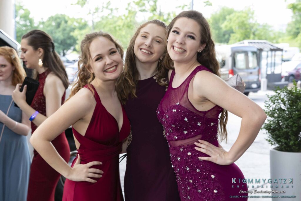 Three people in formal dresses smile for a photo at an outdoor event.