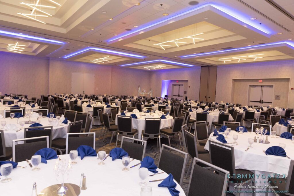 A banquet hall with tables set for an event, featuring white tablecloths, blue napkins, glassware, and chairs. The room is well-lit with modern ceiling lights.