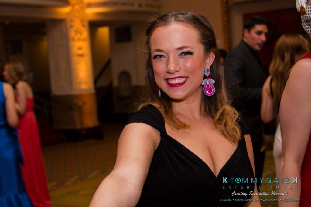 A woman in a black dress smiles at the camera in an indoor event setting. She is wearing colorful earrings and has braided hair.
