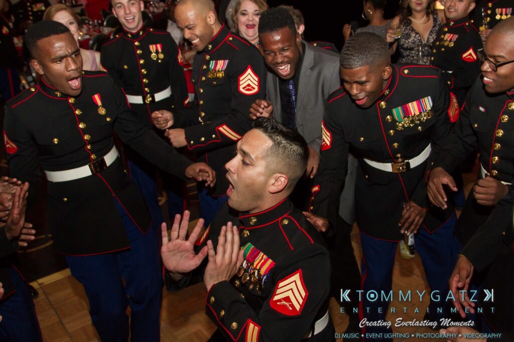 A group of Marines and a man in a suit joyfully celebrate and dance at an event.
