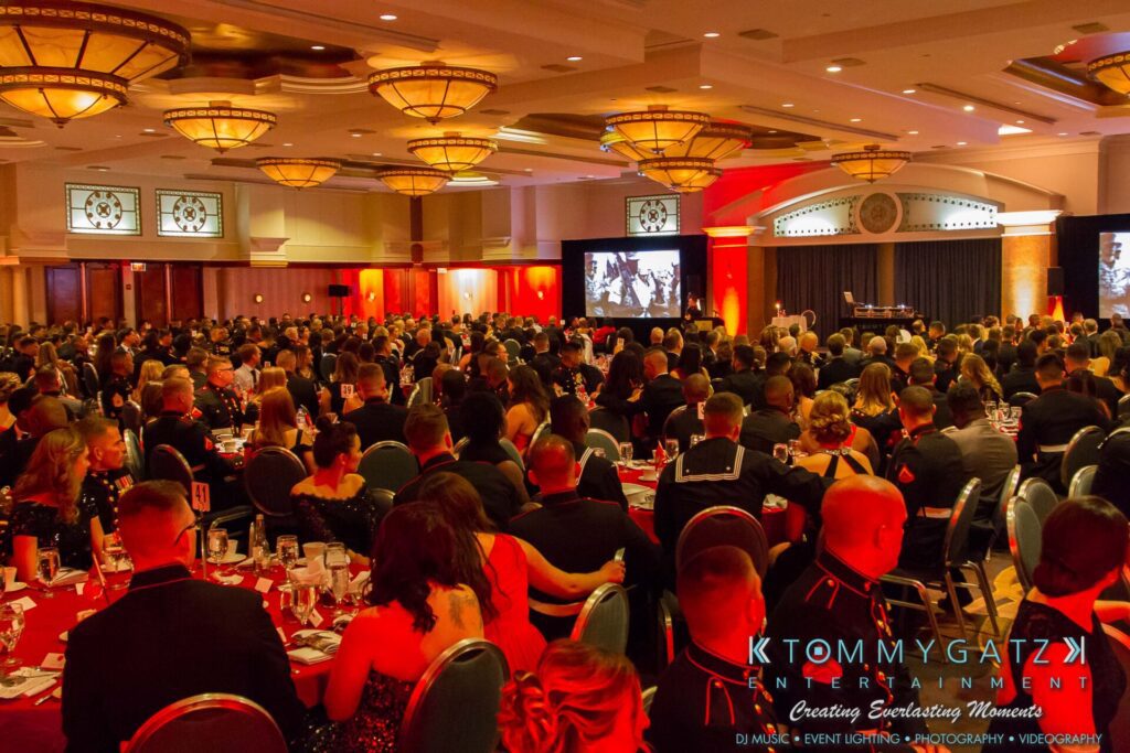 A large, formal event with seated guests in a banquet hall. The room is decorated with chandeliers and red lighting. A screen displays an image at the front.