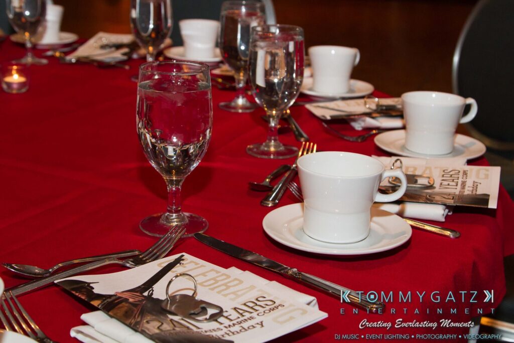 A red tablecloth with neatly arranged dinnerware, including cups, saucers, and glasses. A folded napkin and booklets are placed by each setting.