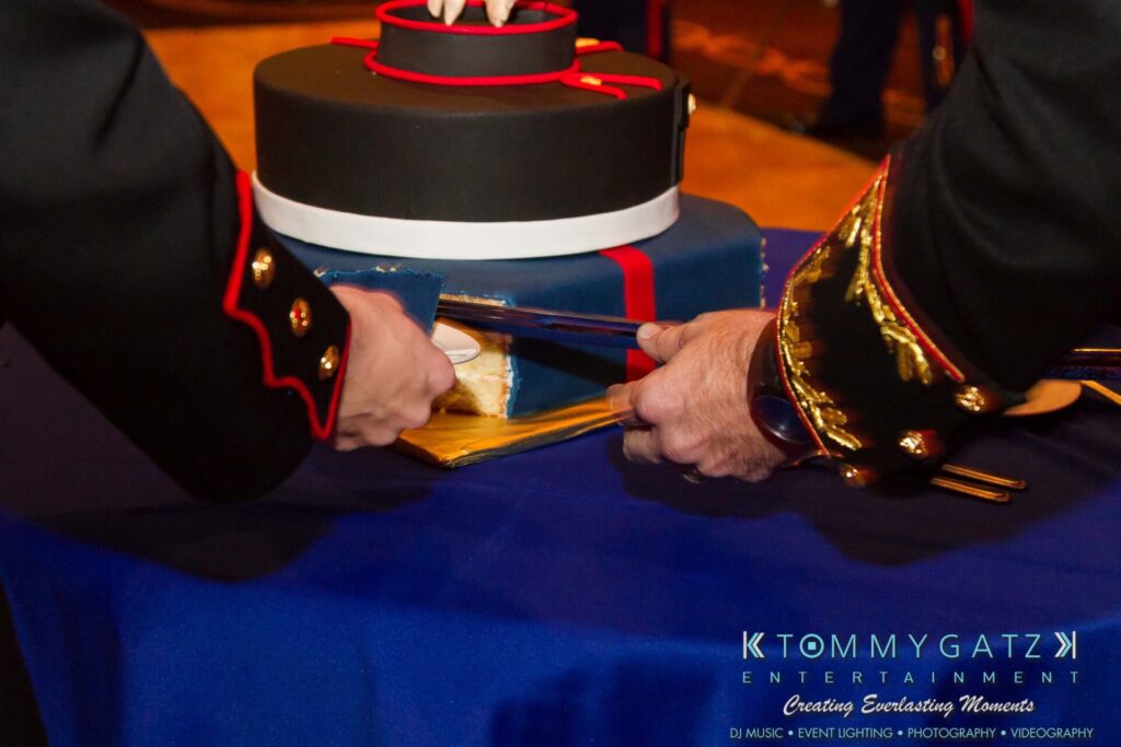 Two uniformed individuals cut a blue and red cake with a ceremonial sword on a blue tablecloth, featuring a logo for Tommy Gatz Entertainment in the bottom right corner.