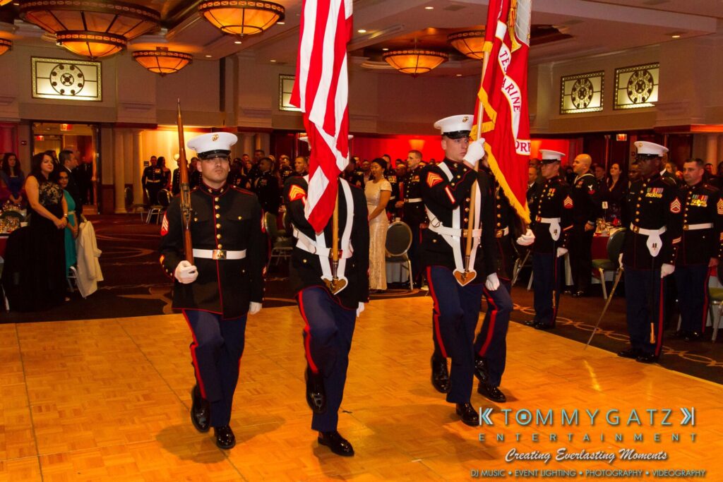 U.S. Marines in dress uniforms perform a color guard ceremony at an indoor event with attendees watching.