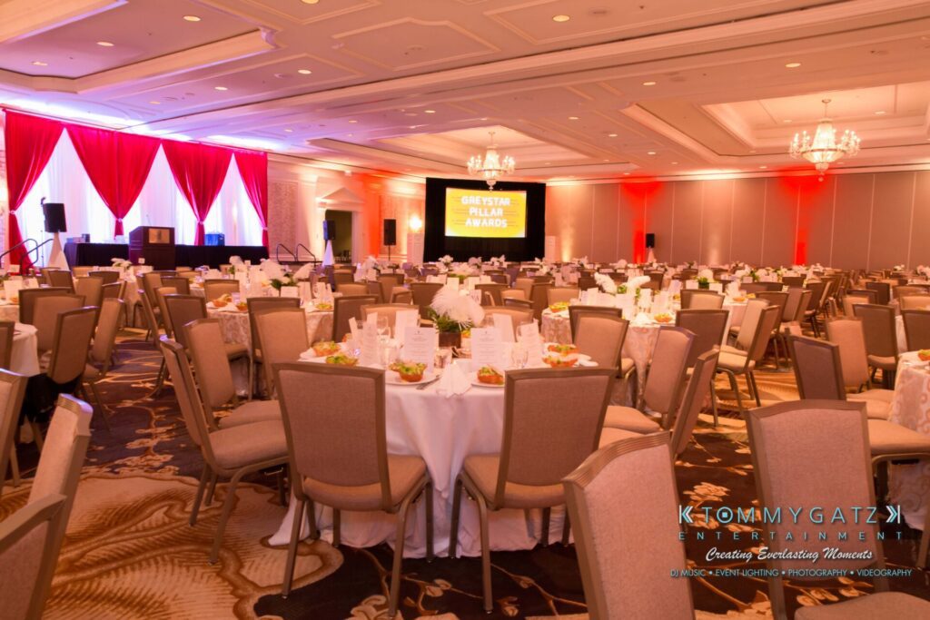 A decorated banquet hall with round tables set for an event. A stage with red curtains and a lit sign is at the front.