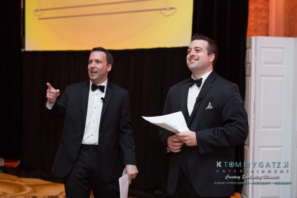 Two men in tuxedos stand on stage, smiling and holding papers. One points while the other looks ahead.