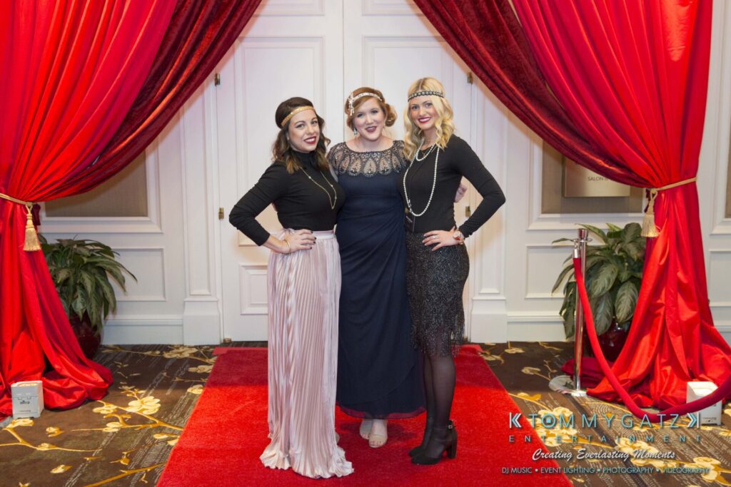 Three women pose on a red carpet, dressed in vintage-style outfits with headbands and pearl necklaces. Red curtains and a roped-off area are in the background.