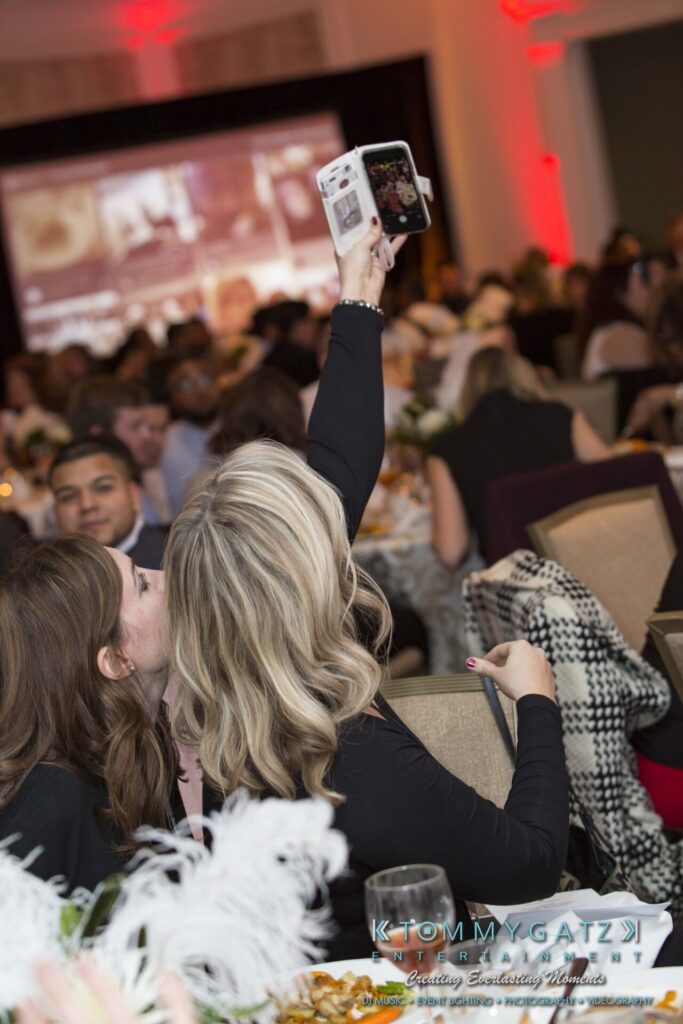 Two people seated at a table taking a selfie with a smartphone at an indoor event. The room has a crowd and red lighting.