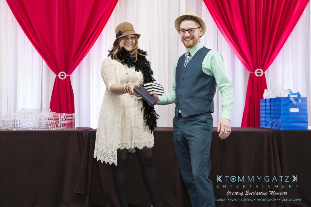 Two people stand in front of a table with red and white drapes. One hands a small stack of books to the other. They are smiling and dressed formally.