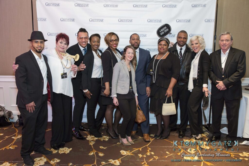 A group of people posing together in front of a branded backdrop at an event. Some are holding props, including a trophy and a sign. They are dressed in business and casual attire.