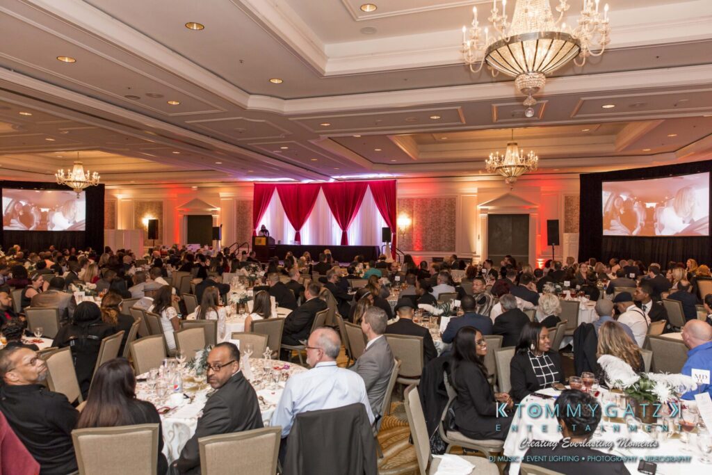 A large conference room filled with seated attendees facing a stage with red curtains and large screens.