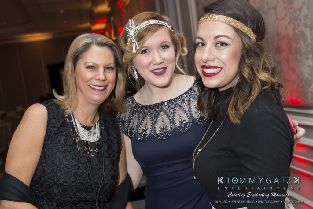 Three people smiling at a formal event, with two wearing headbands and evening dresses. A logo in the corner reads "Tommy Gatz Entertainment.
