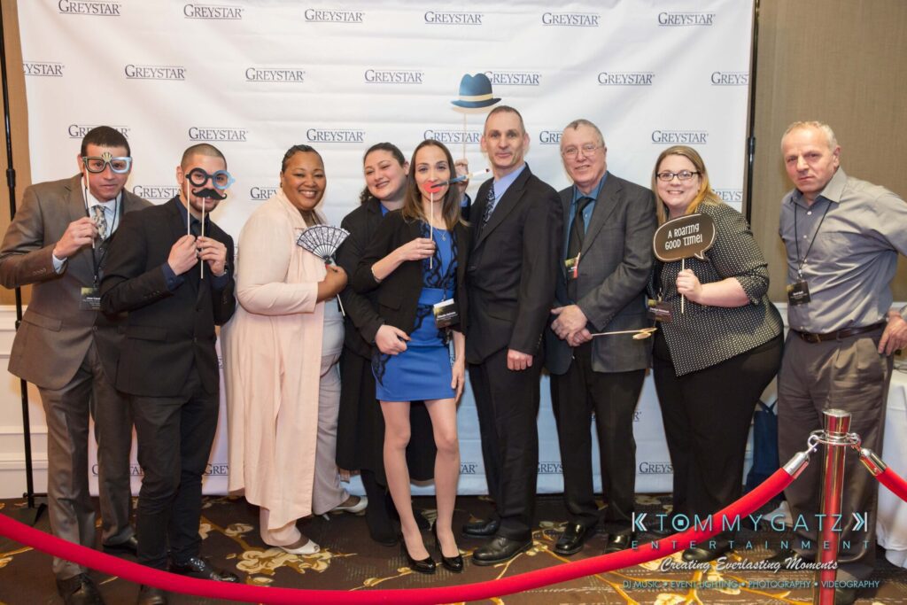 A group of people posing with props in front of a backdrop that reads "GREYSTAR," standing on a carpeted floor with a red velvet rope barrier.
