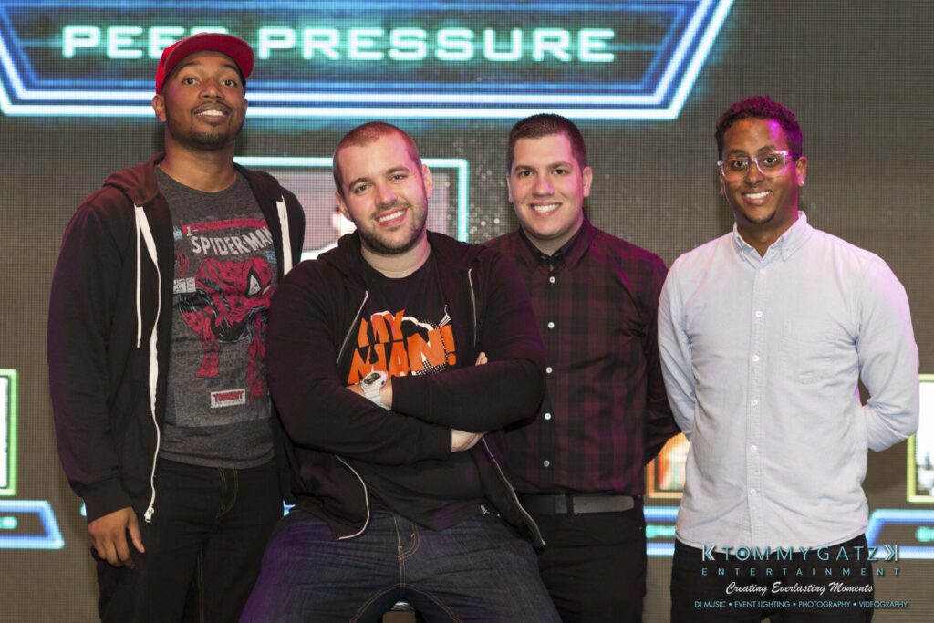 Four men standing and posing for a photo indoors, smiling, with a sign that reads "Peer Pressure" in the background.
