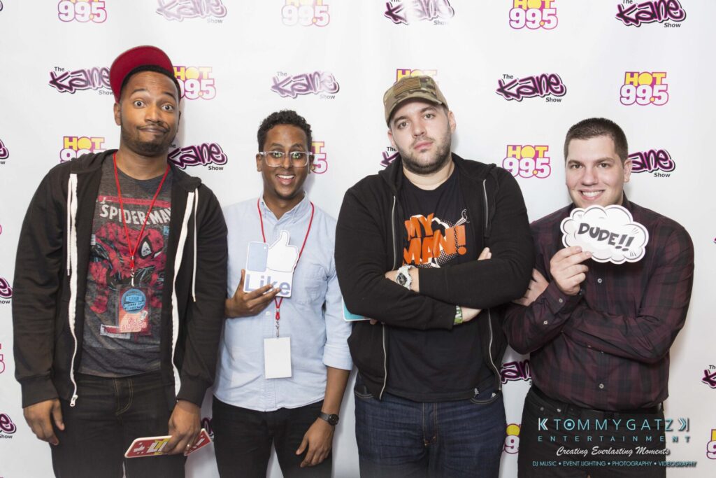 Four people pose in front of a backdrop with "Hot 99.5" logos, holding props and smiling.