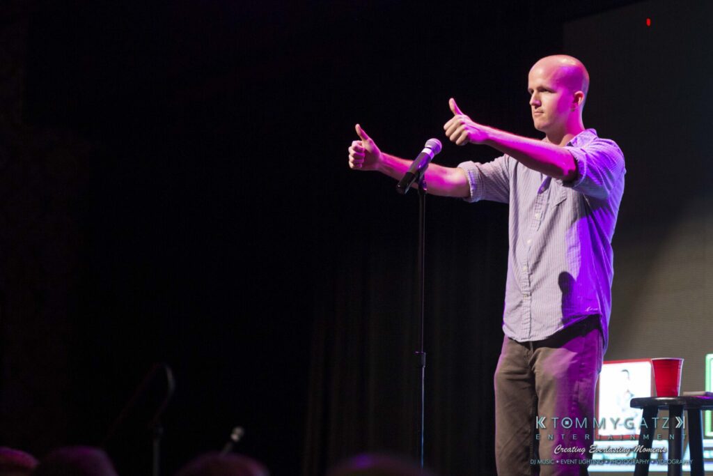 A man stands on stage performing with a microphone, giving two thumbs up.