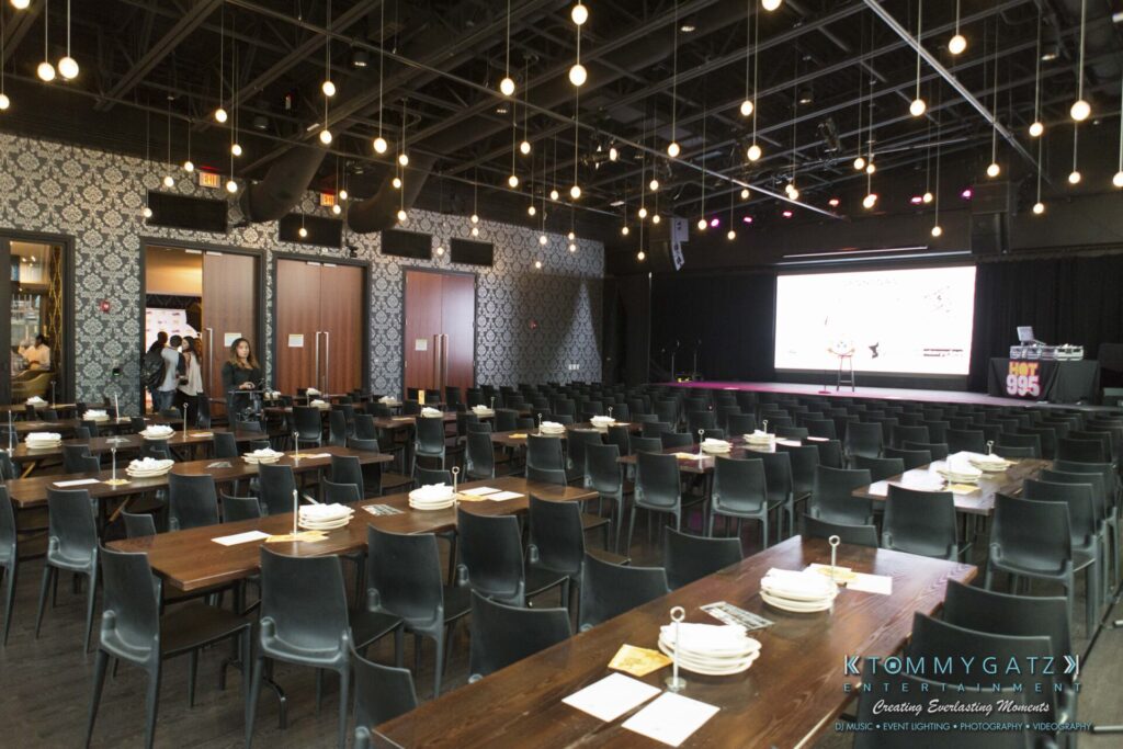 A dimly lit event space with tables set for a meal, plates and utensils neatly arranged. Rows of chairs face a stage with a large screen displaying white and purple graphics.