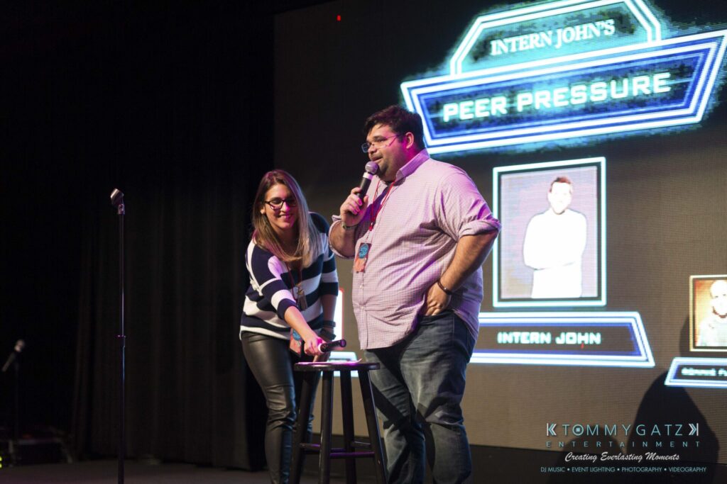 Two people stand on stage with microphones in front of a sign that reads "Intern John's Peer Pressure." They are near a small round table.