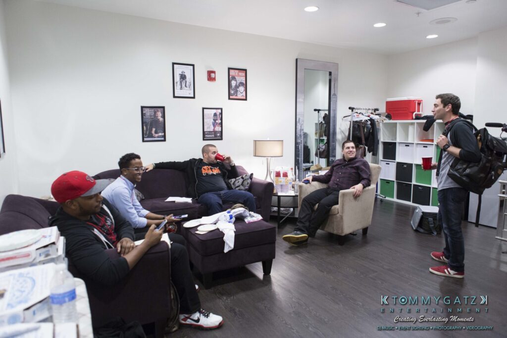 A group of people casually sitting and talking in a living room, with a mix of couches and chairs, papers, and a pizza box visible.