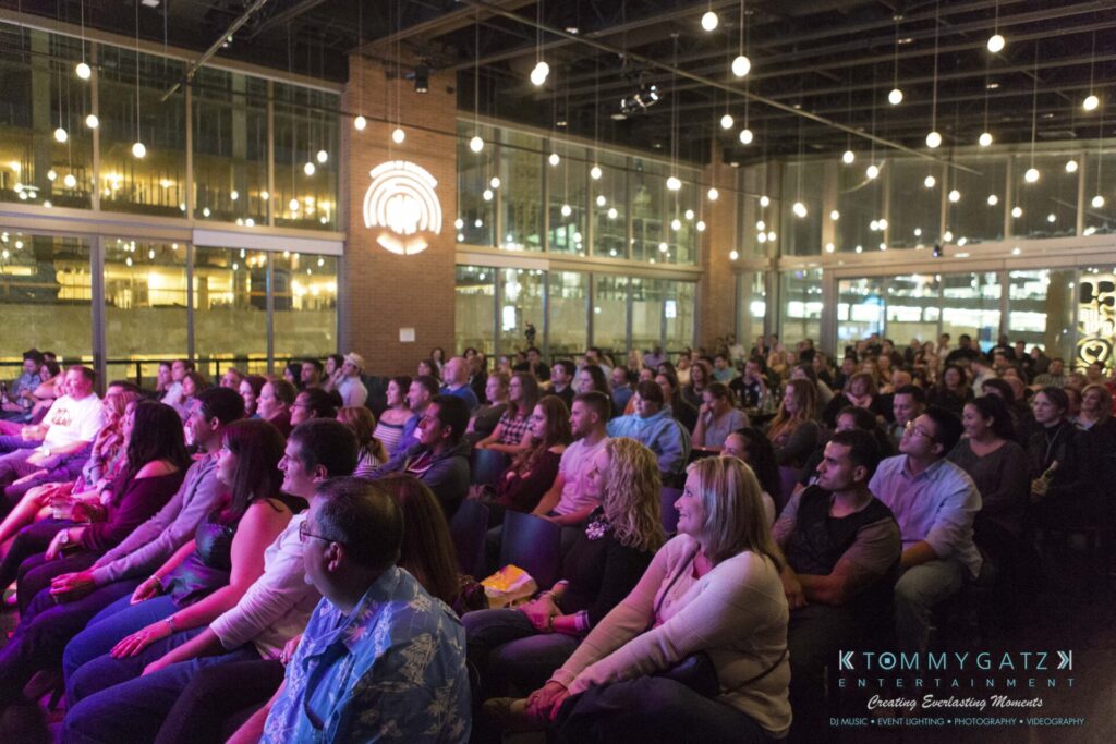 Audience seated at an indoor event, attentively watching a stage performance, with string lights hanging from the ceiling and large windows in the background.