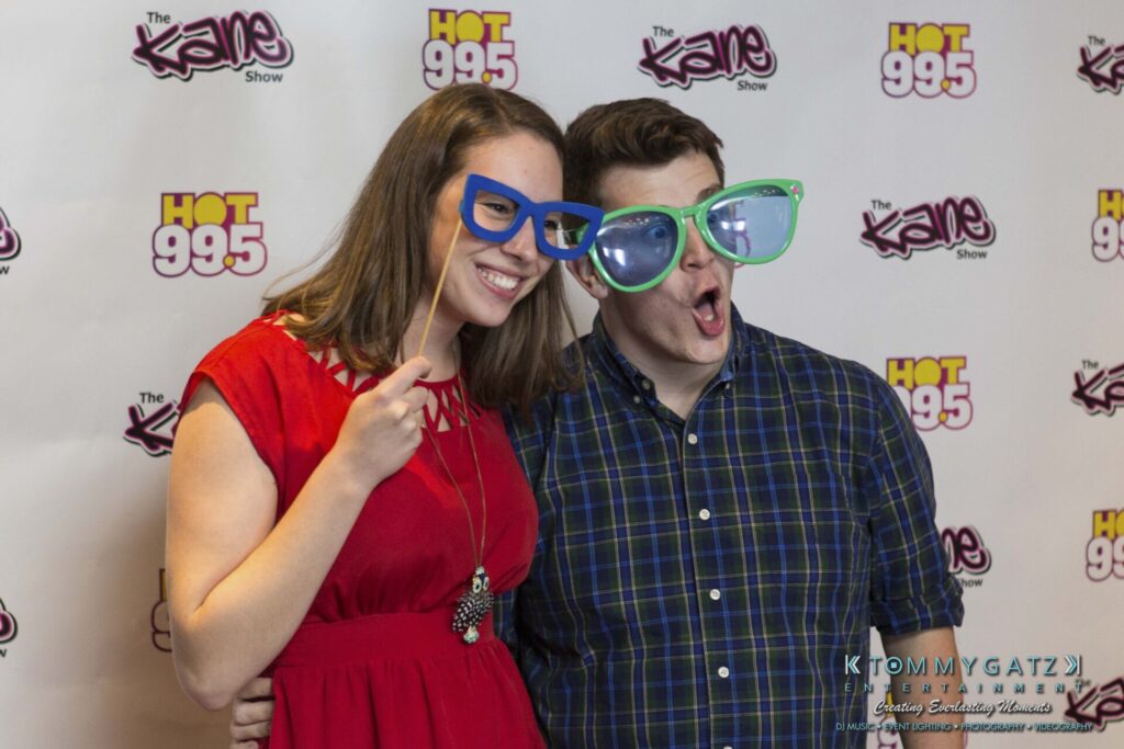 Two people pose playfully with oversized glasses in front of a step and repeat banner featuring "The Kane Show" and "HOT 99.5" logos.