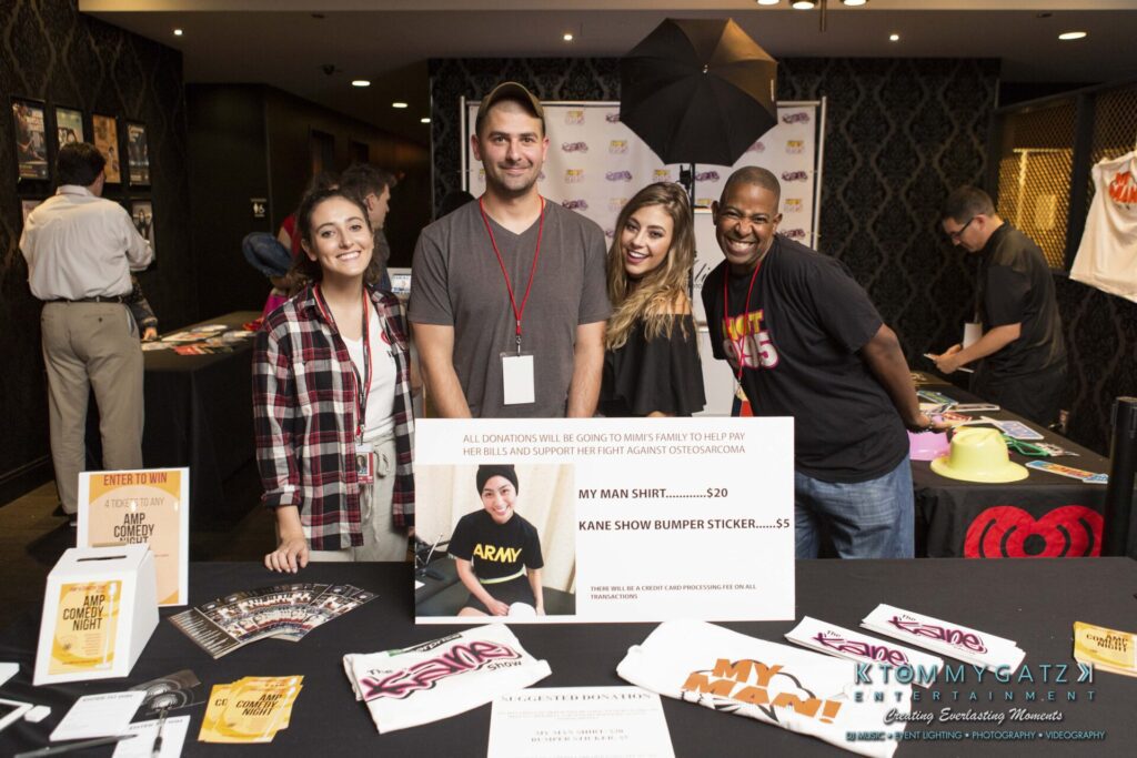 Four people stand behind a merchandise table displaying T-shirts, stickers, and a donation sign for charity in a lobby setting.