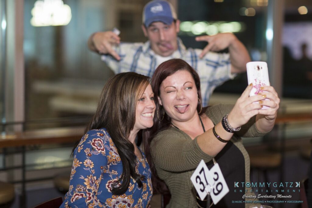 Two women are taking a selfie at a table with a 37.9 sign, while a man in the background makes a playful face and gesture.