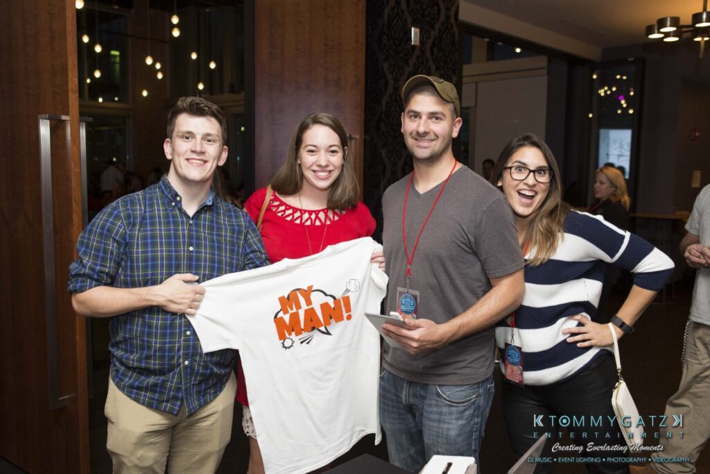 Four people smiling at the camera; one holds a T-shirt with "MY MAN!" printed on it. They stand indoors at an event, wearing casual clothing and event lanyards.