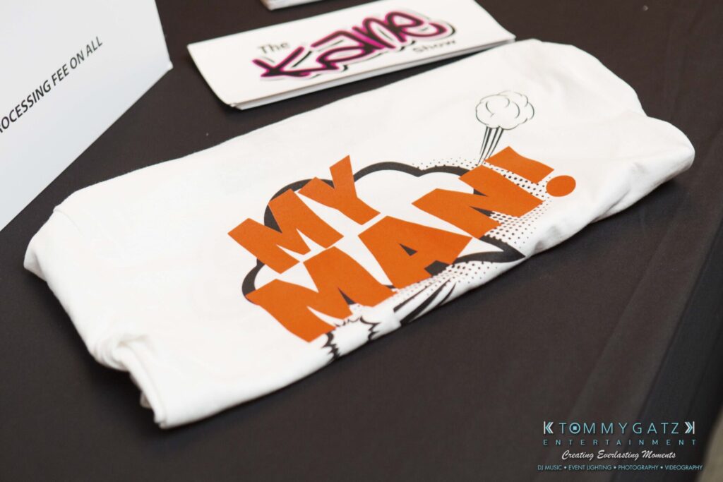 A folded white shirt with "My Man!" printed in bold orange and black letters lies on a table near promotional cards.