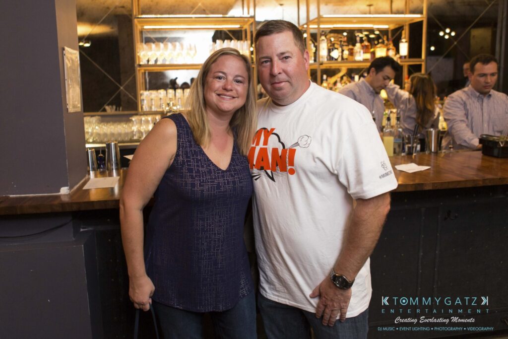 A man and woman stand together in front of a bar, smiling at the camera. Other people are visible in the background.