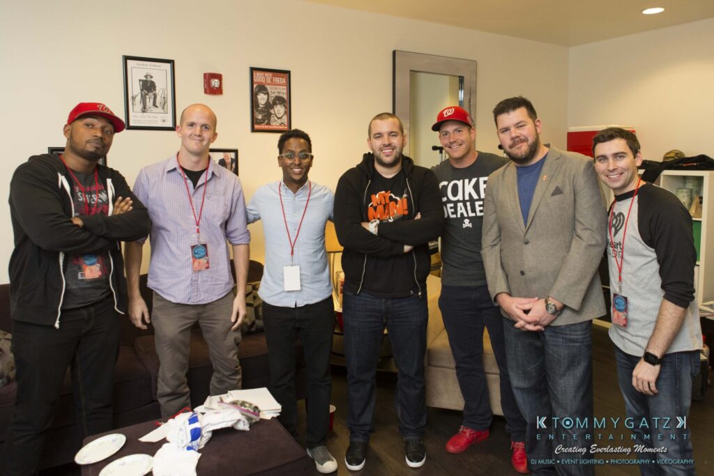 A group of seven men standing indoors, some wearing casual clothing and hats, posing for a photo in a room with framed pictures on the wall.