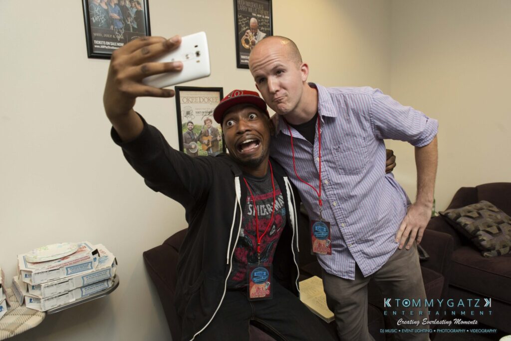 Two men taking a selfie in a room with pizza boxes on the side table. One is wearing a black jacket and red cap, and the other is wearing a striped shirt. Posters are on the wall behind them.