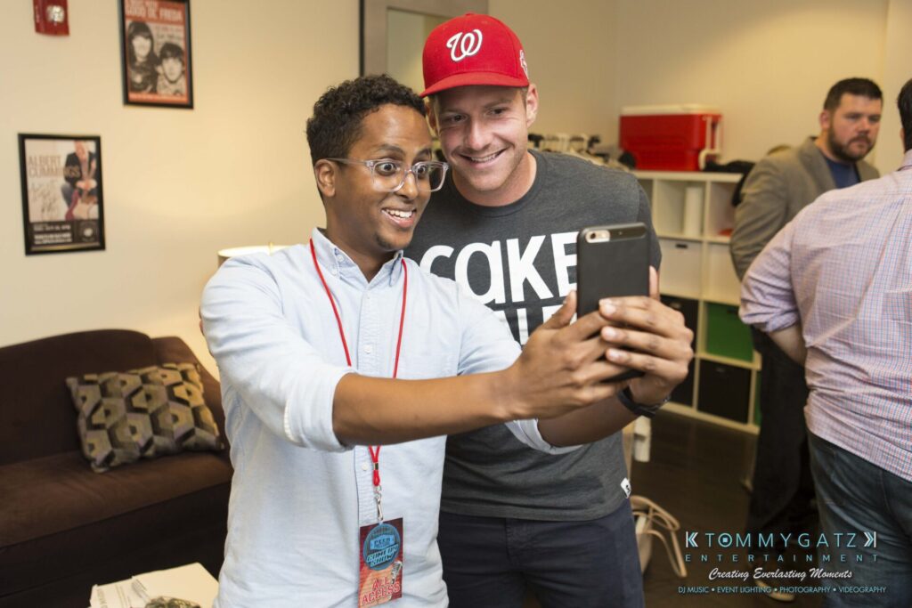 Two smiling men taking a selfie together in a room with casual decor. One is wearing a red cap and gray shirt, the other has glasses and a lanyard.