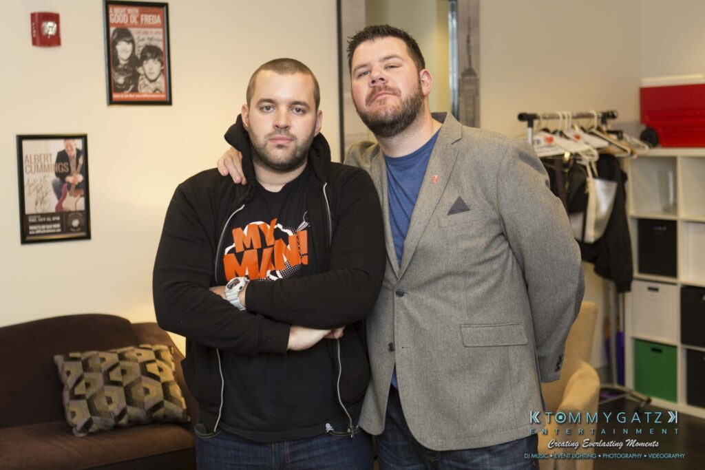 Two men stand together in a room, one with arms crossed. The room has posters, a couch with cushions, and a clothing rack.