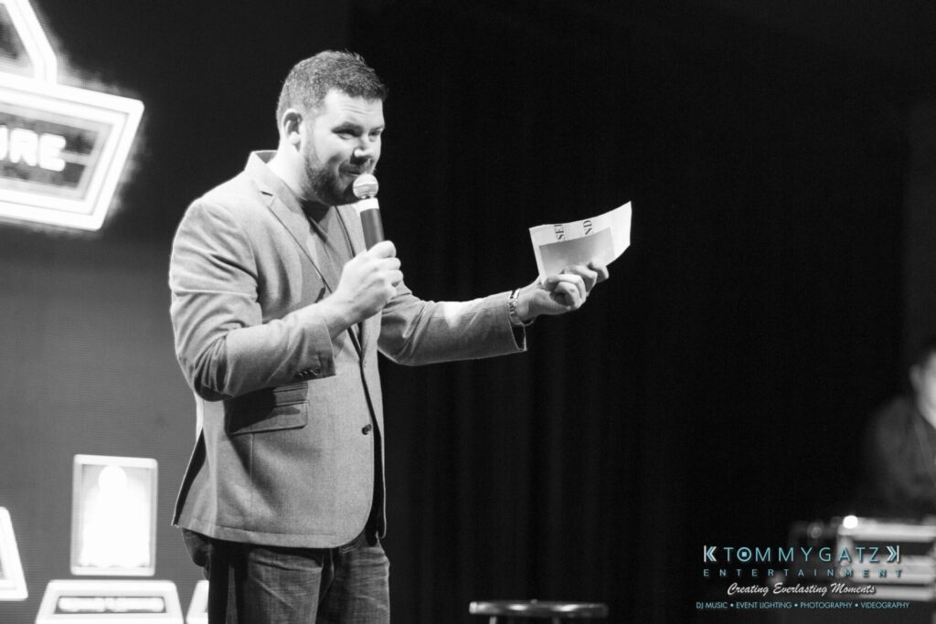Man holding a microphone and papers, speaking on stage in a black and white setting.