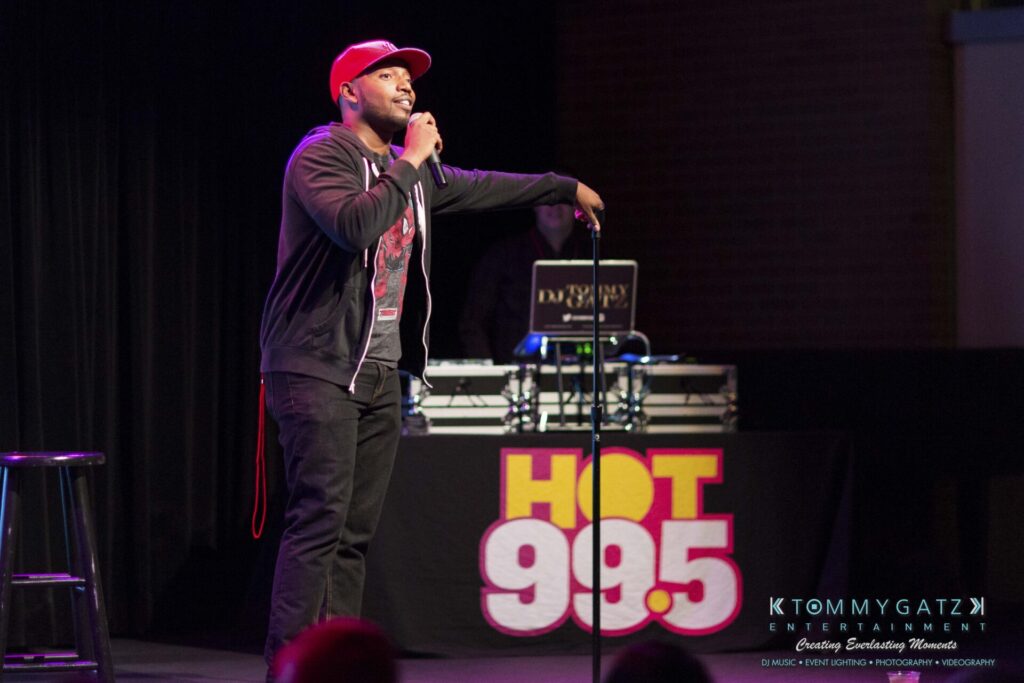 A person on stage performing stand-up comedy, holding a microphone. A DJ booth with "HOT 99.5" is visible in the background.