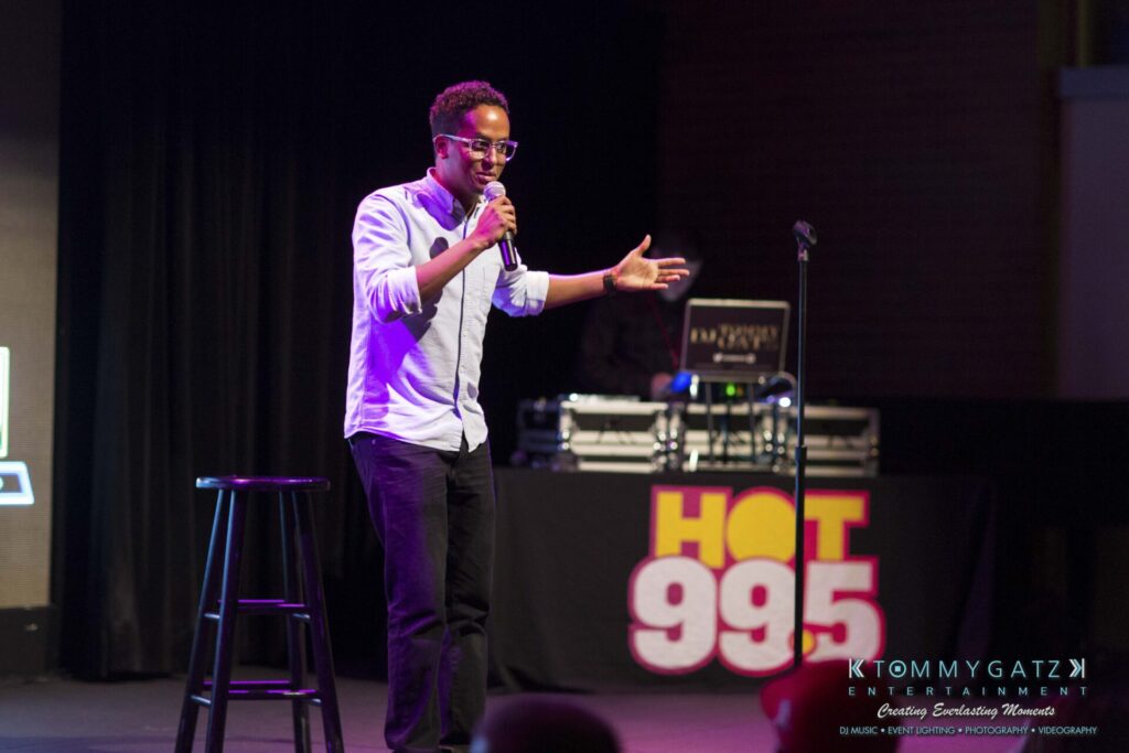 A person performs stand-up comedy on stage with a microphone and a stool. A DJ booth with "Hot 99.5" is visible in the background.